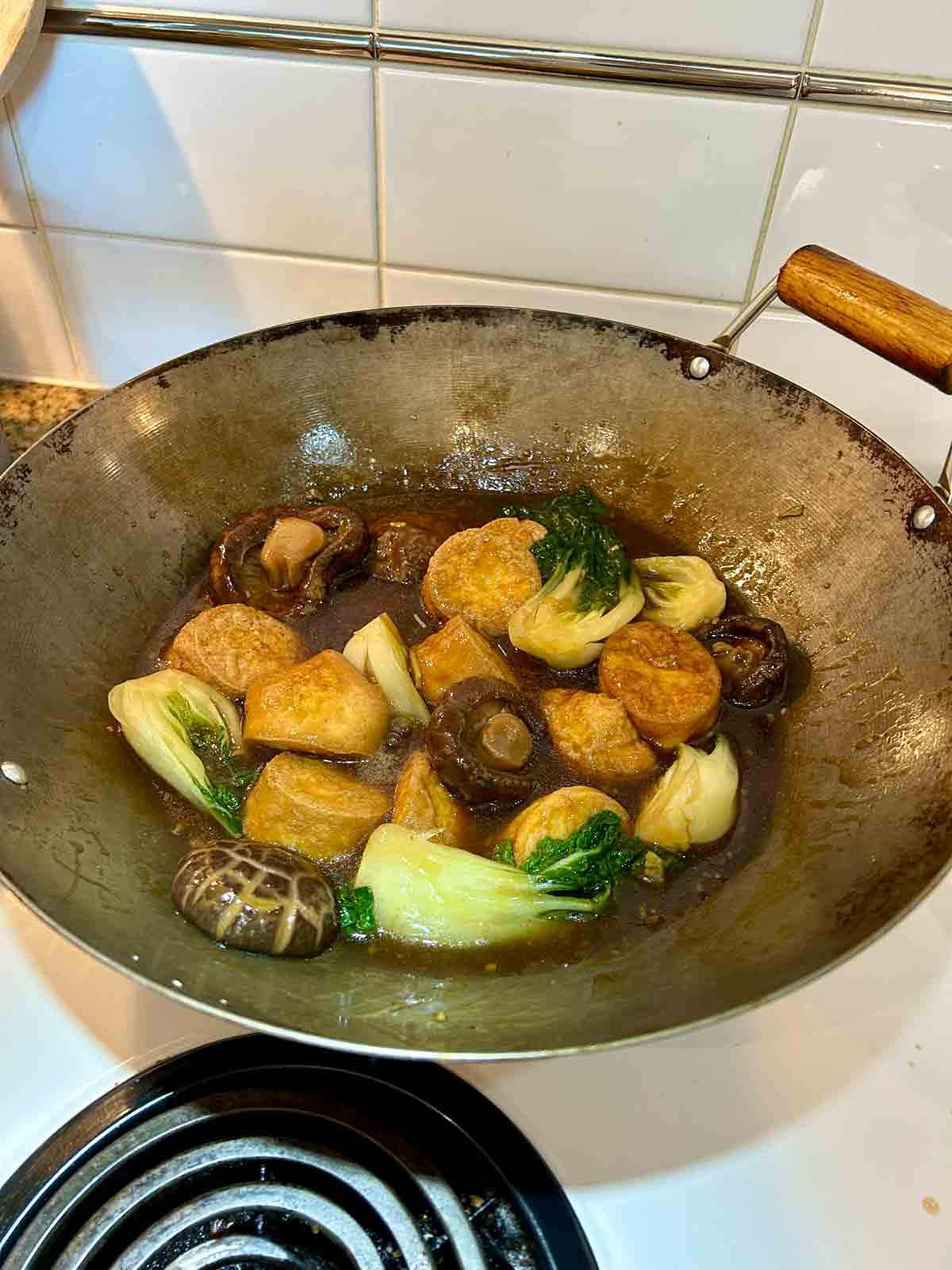 egg tofu, shiitake mushrooms, and bok choy being tossed with brown sauce in wok.