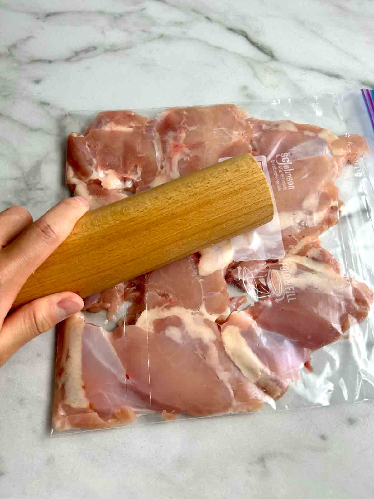 chicken thighs being flattened with rolling pin in zipper-lock bag.