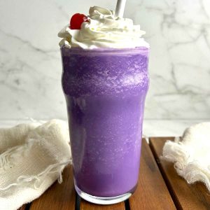 ube milkshake in tall glass with whipped cream and cherry on top.