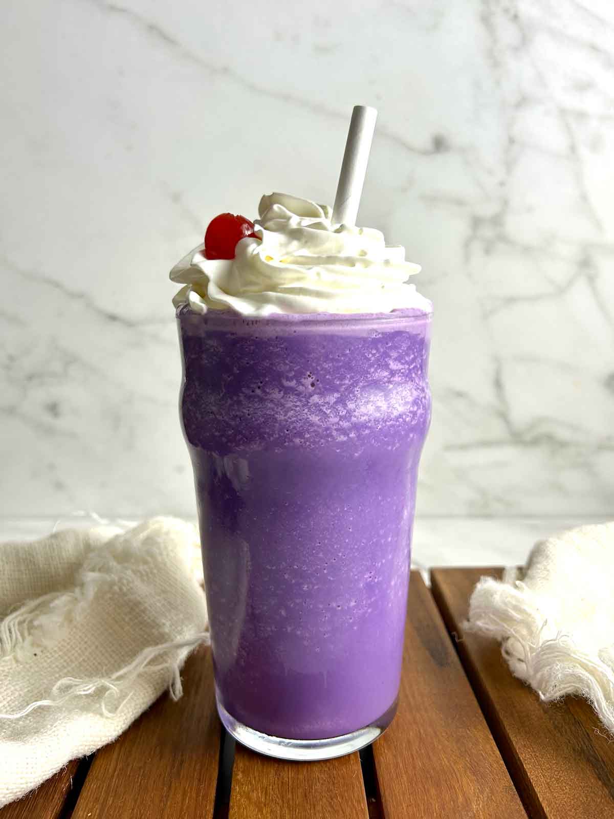 ube milkshake in tall glass with whipped cream and cherry on top.