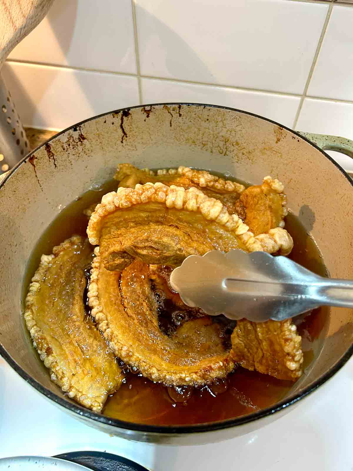 tongs pulling out fried chicharron with puffy skin from hot oil.