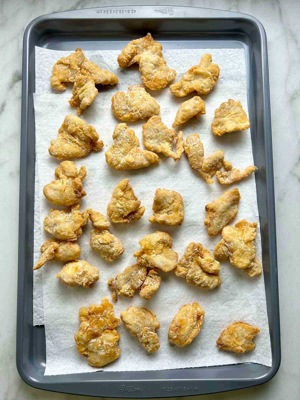 fried chicken pieces drying on paper towel lined baking sheet.