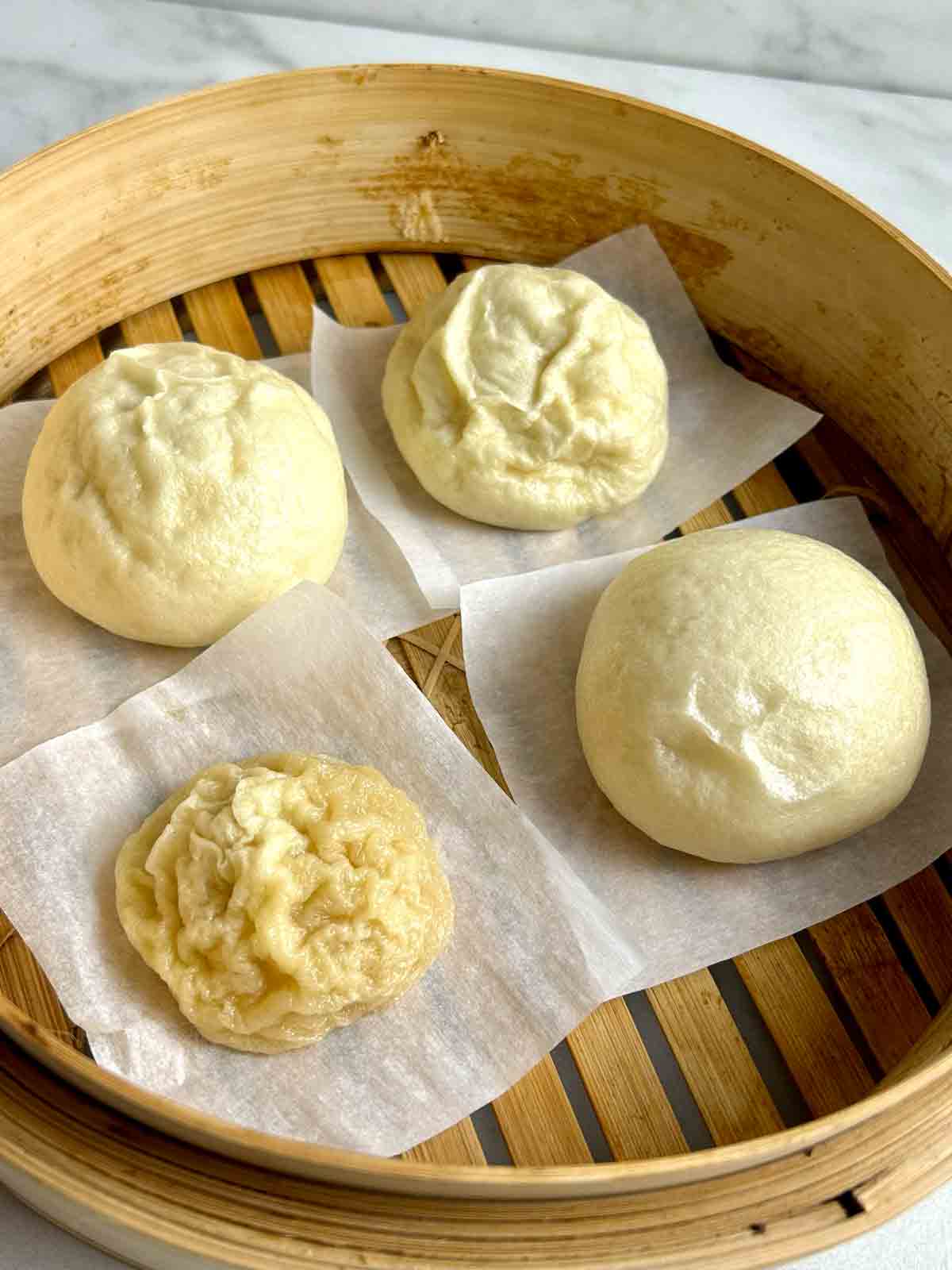 four steamed buns in a bamboo steamer with varying results: one fully collapsed, one slightly wrinkled, another heavily wrinkled, and one that appears normal.