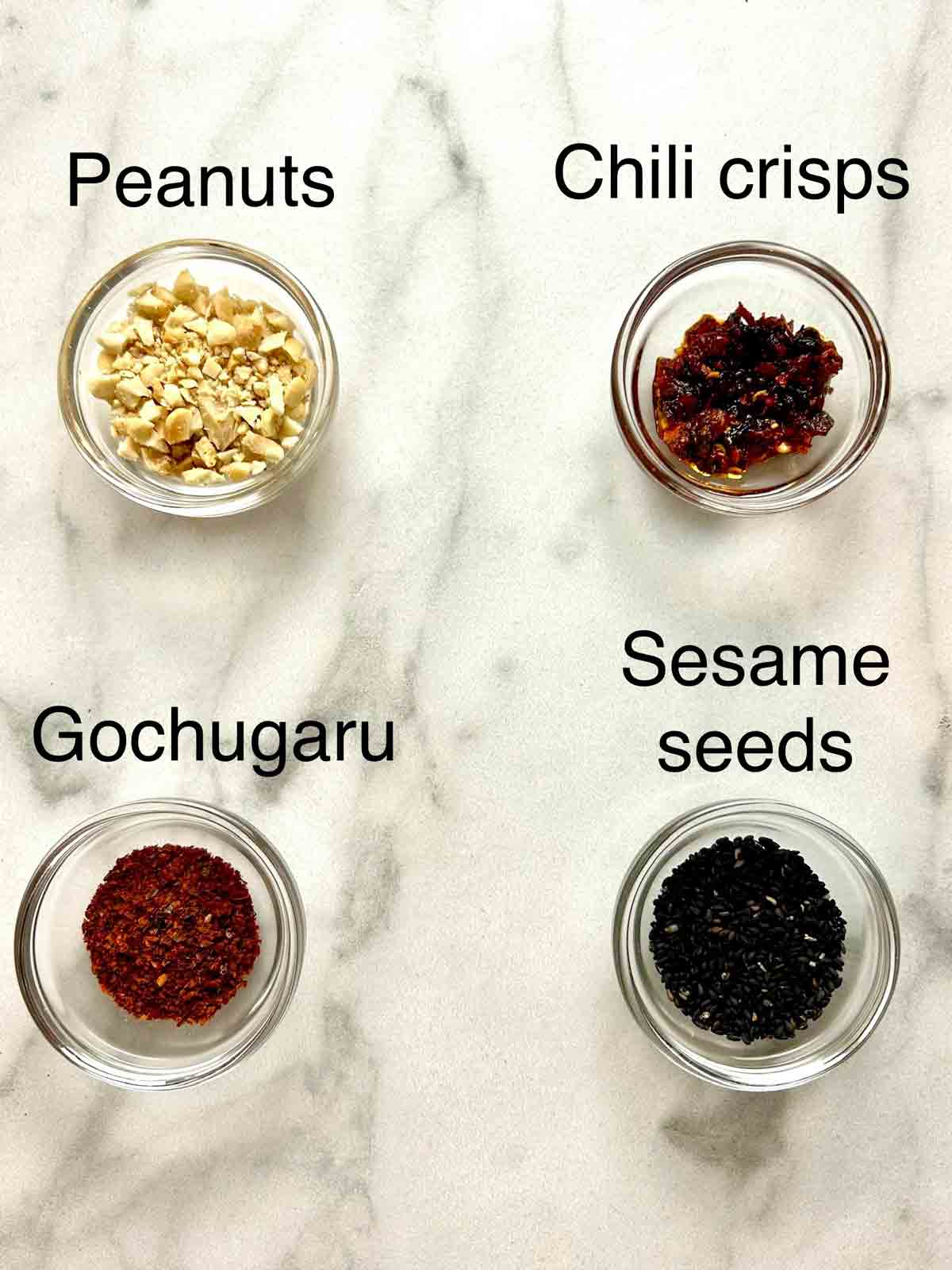 Four separate bowls containing chopped peanuts, chili crisps, gochugaru, and sesame seeds.