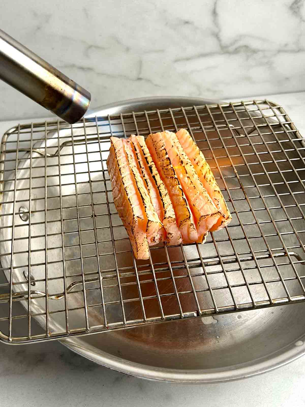 salmon slices being torched with blowtorch on wire rack.