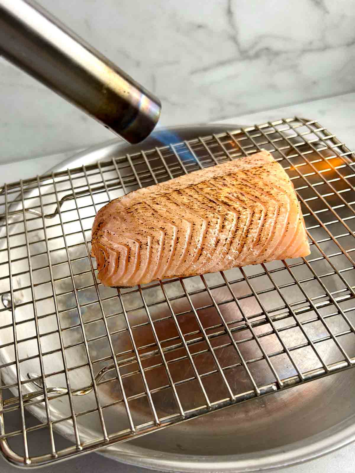 unsliced salmon fillet being torched with blowtorch on wire rack.