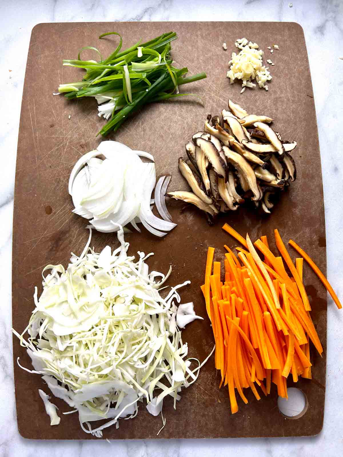 sliced scallions, minced garlic, sliced onions, sliced shiitake mushrooms, shredded cabbage, and julienned carrots on cutting board.