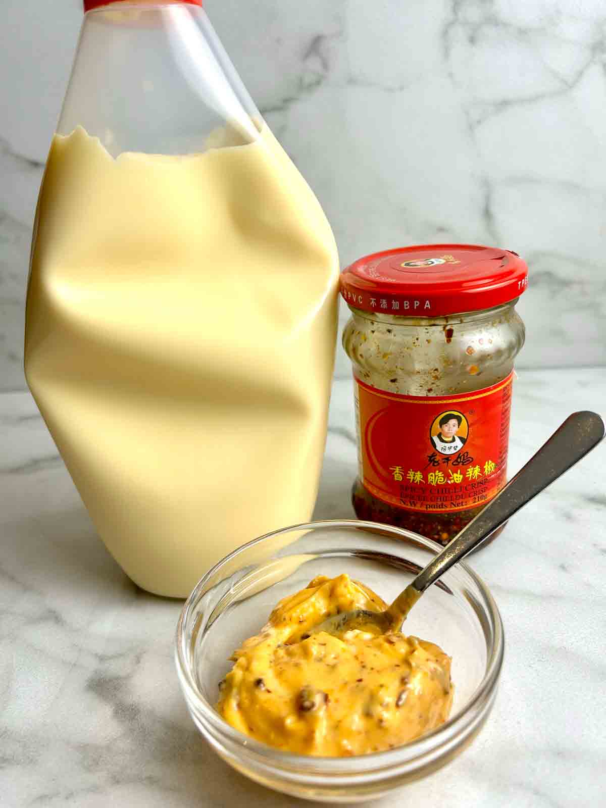 bowl of chili crisp mayo with bottle of mayo and jar of chili oil in background.