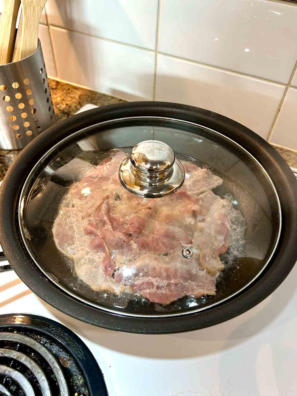 bacon cooking in pan with water and lid covering it to trap steam.