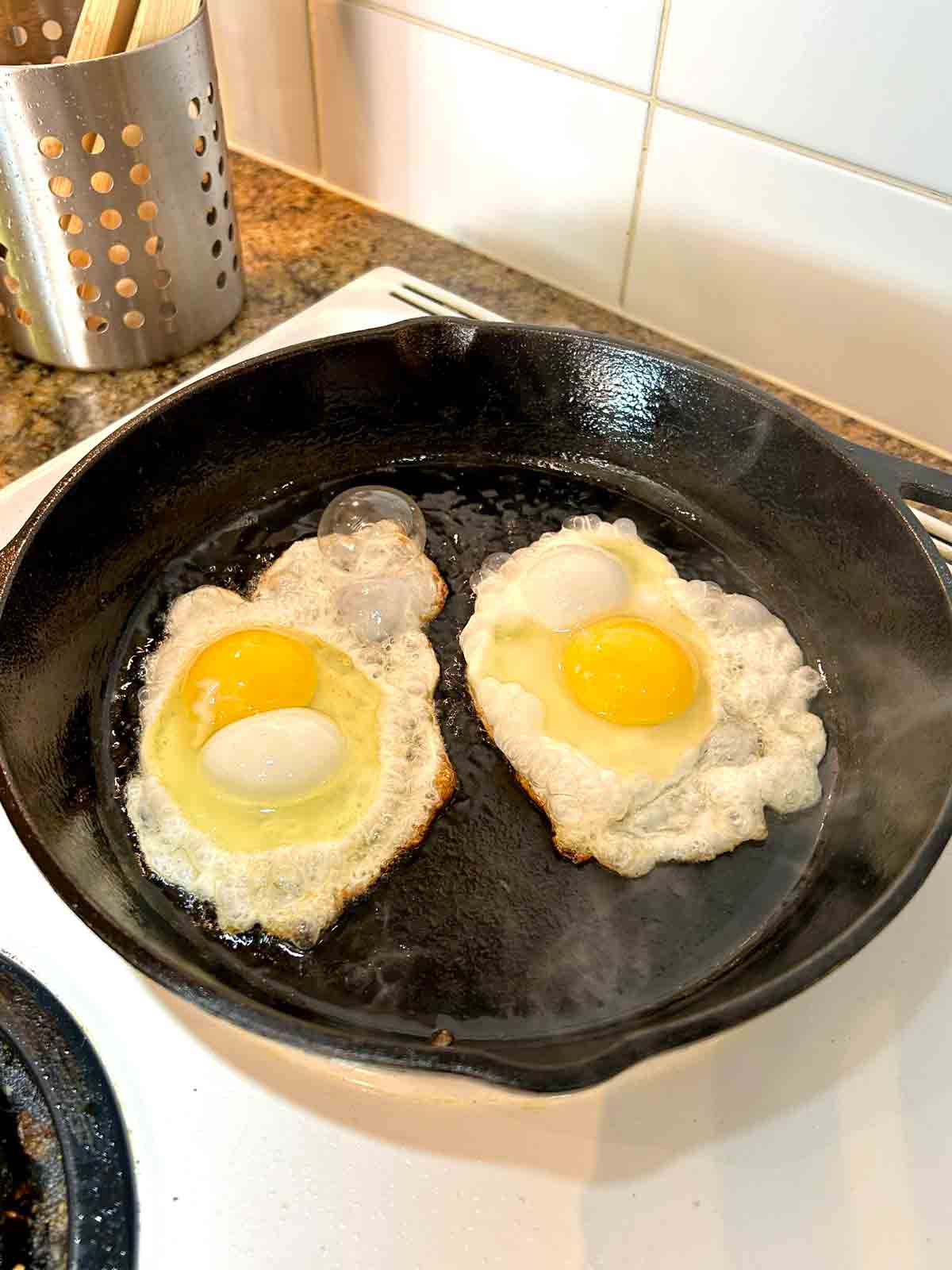 two eggs being fried in hot cast iron pan.