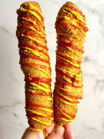 hand holding two Korean corn dogs covered with ketchup and mustard.