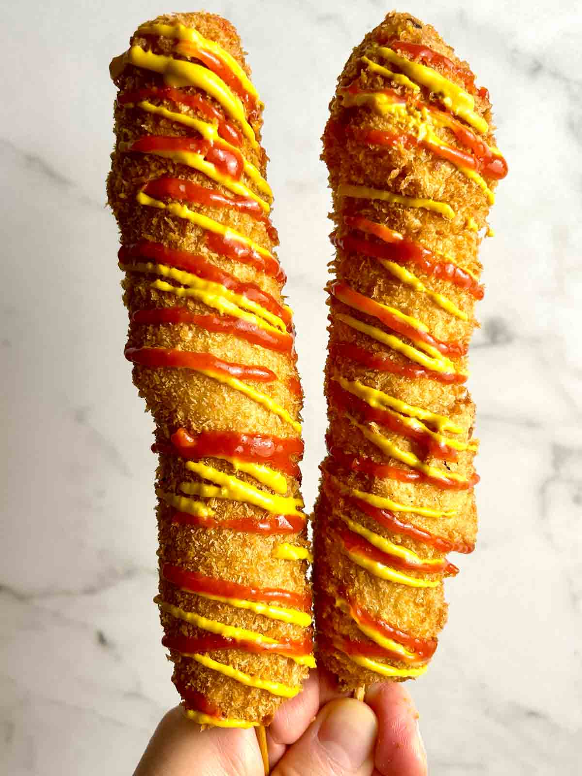 hand holding two Korean corn dogs covered with ketchup and mustard.