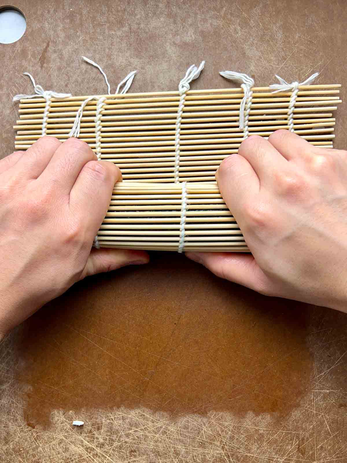 squeezing kimbap inside bamboo mat with half of bamboo mat hanging off the side.