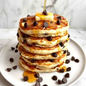 stack of chocolate chip pancakes on plate; maple syrup being drizzled on top.