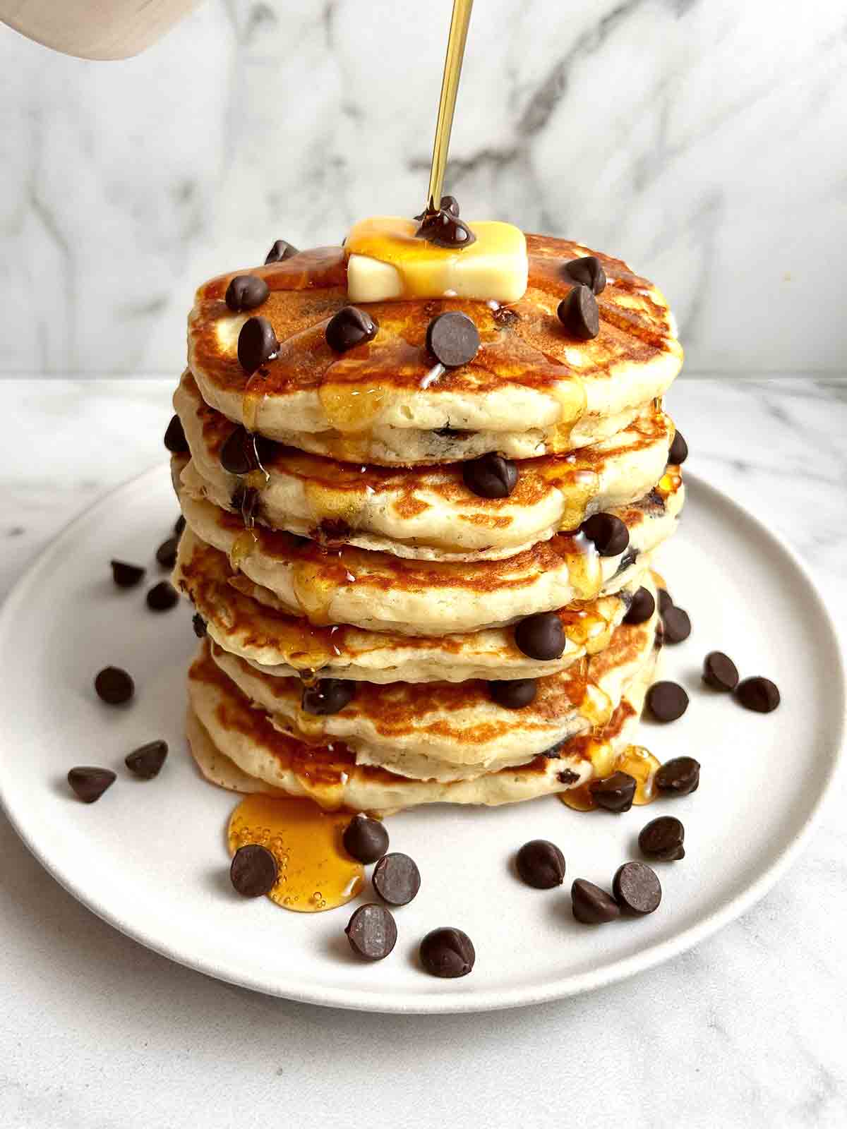 stack of chocolate chip pancakes on plate; maple syrup being drizzled on top.