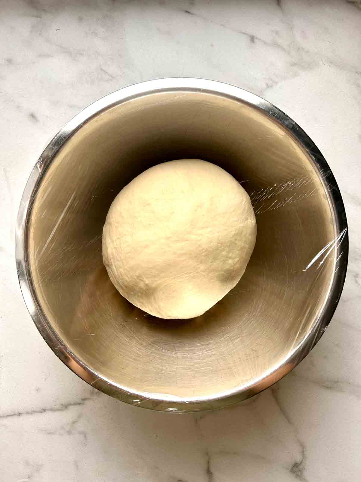 kneaded dough resting in bowl with plastic wrap covering it.