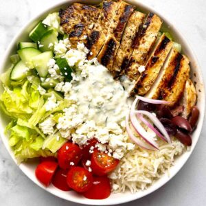 Greek chicken bowl with lettuce, cucumber, cherry tomatoes, tzatziki, red onion, olives, feta cheese, and grilled chicken.