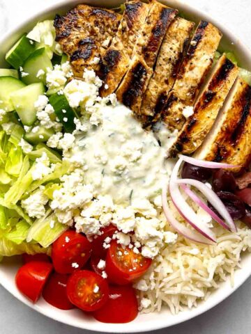 Greek chicken bowl with lettuce, cucumber, cherry tomatoes, tzatziki, red onion, olives, feta cheese, and grilled chicken.