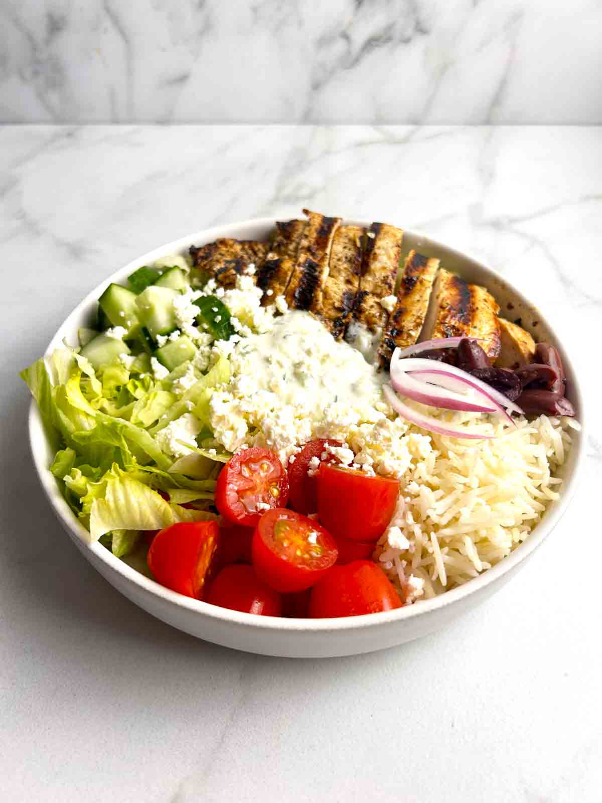 Greek chicken bowl with lettuce, cucumber, cherry tomatoes, tzatziki, red onion, olives, feta cheese, and grilled chicken.