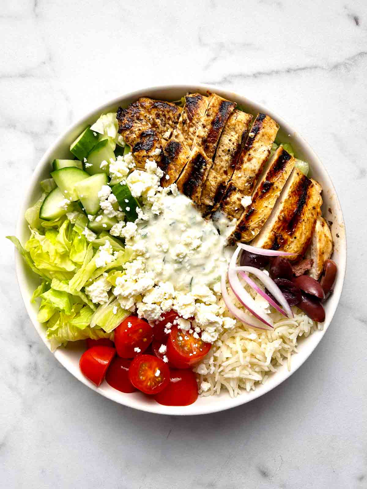 Greek chicken bowl with lettuce, cucumber, cherry tomatoes, tzatziki, red onion, olives, feta cheese, and grilled chicken.
