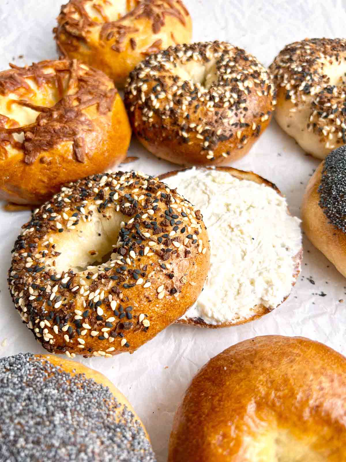 assorted sourdough bagels on parchment paper with one cut in half with cream cheese.