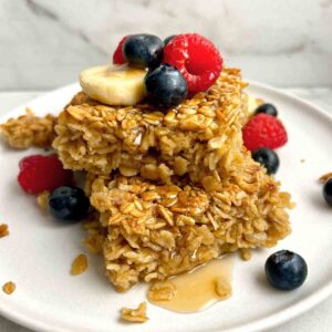 two slices of baked oatmeal on top of each other; maple syrup drizzled on top and fresh berries on plate.