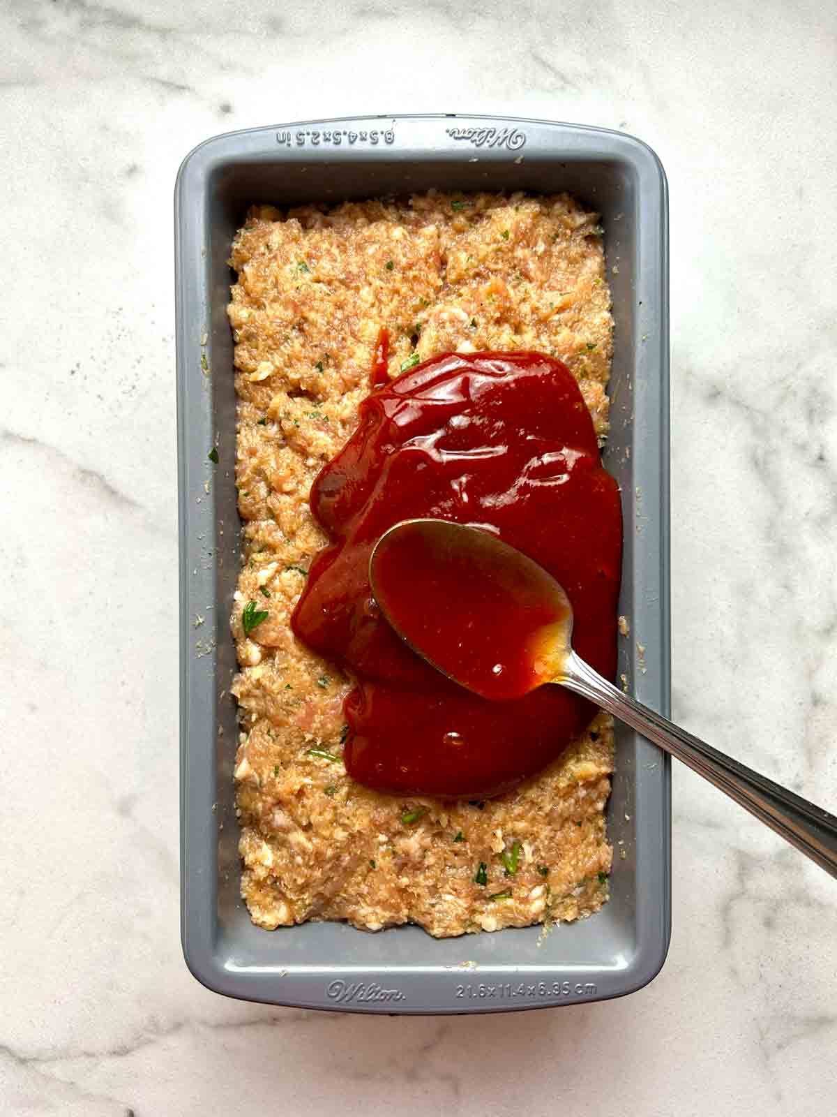 ketchup being poured on meat mixture in loaf pan.