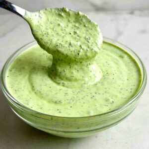 close up of spoon lifting green goddess dressing from glass bowl.