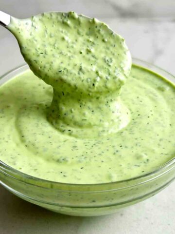 close up of spoon lifting green goddess dressing from glass bowl.
