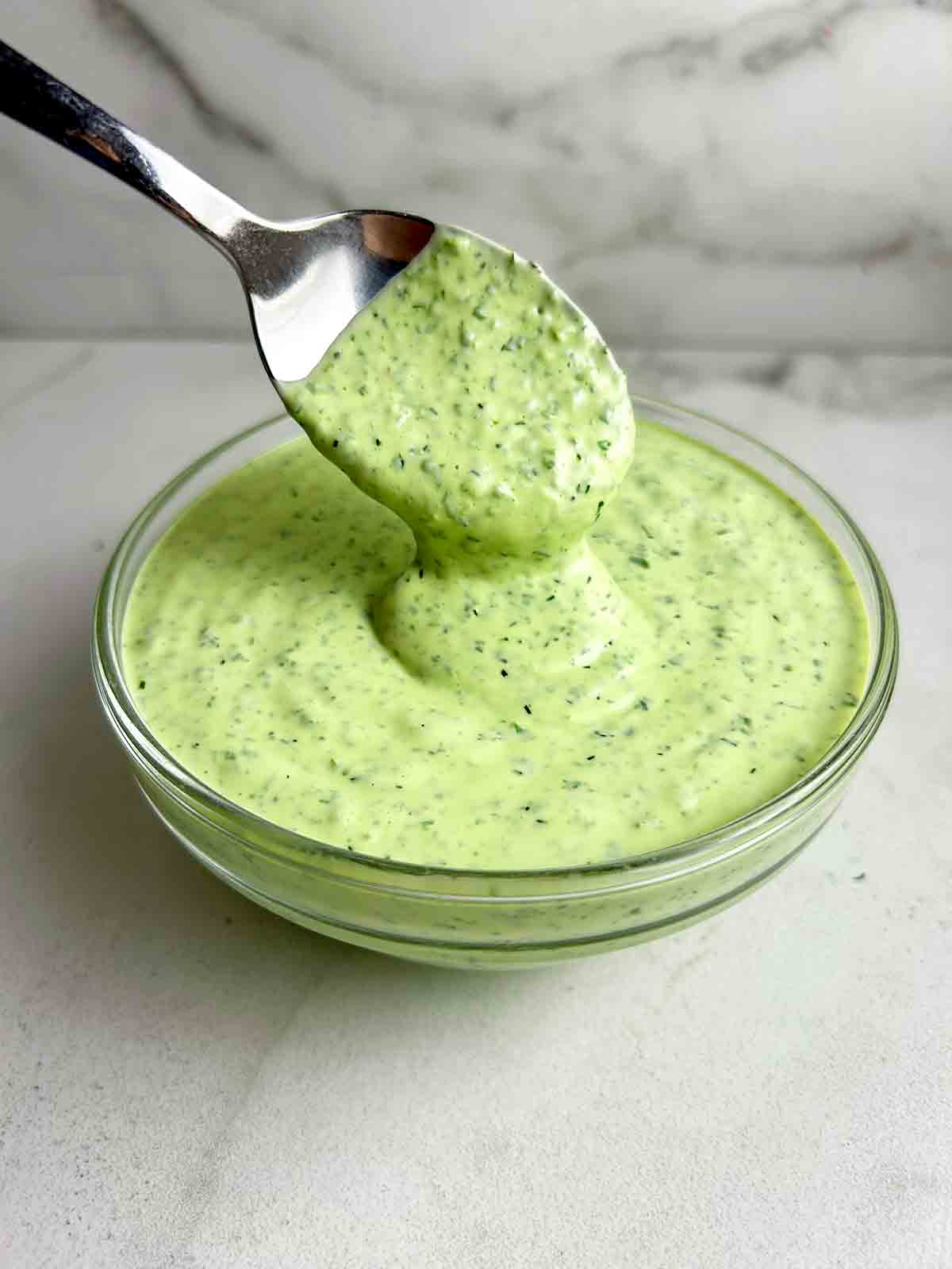 close up of spoon lifting green goddess dressing from glass bowl.
