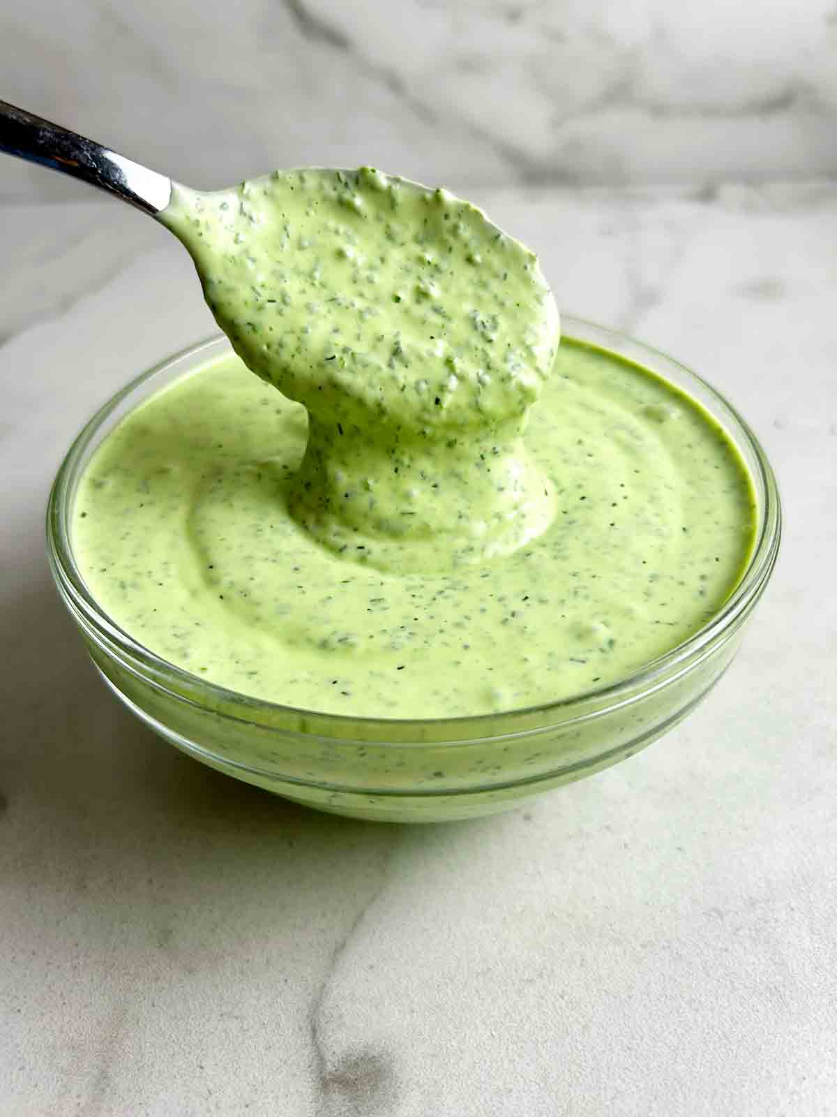 close up of spoon lifting green goddess dressing from glass bowl.