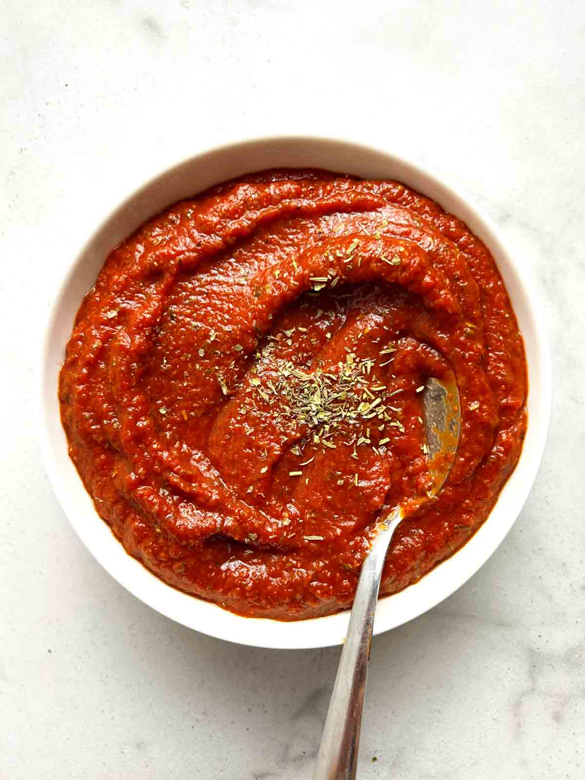 bowl of pizza sauce with dried herbs sprinkled on top and spoon in it.