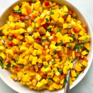 A bowl of mango salsa with diced mango, red onion, and cilantro, mixed with red bell pepper. A spoon rests in the bowl.