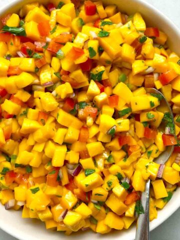 A bowl of mango salsa with diced mango, red onion, and cilantro, mixed with red bell pepper. A spoon rests in the bowl.