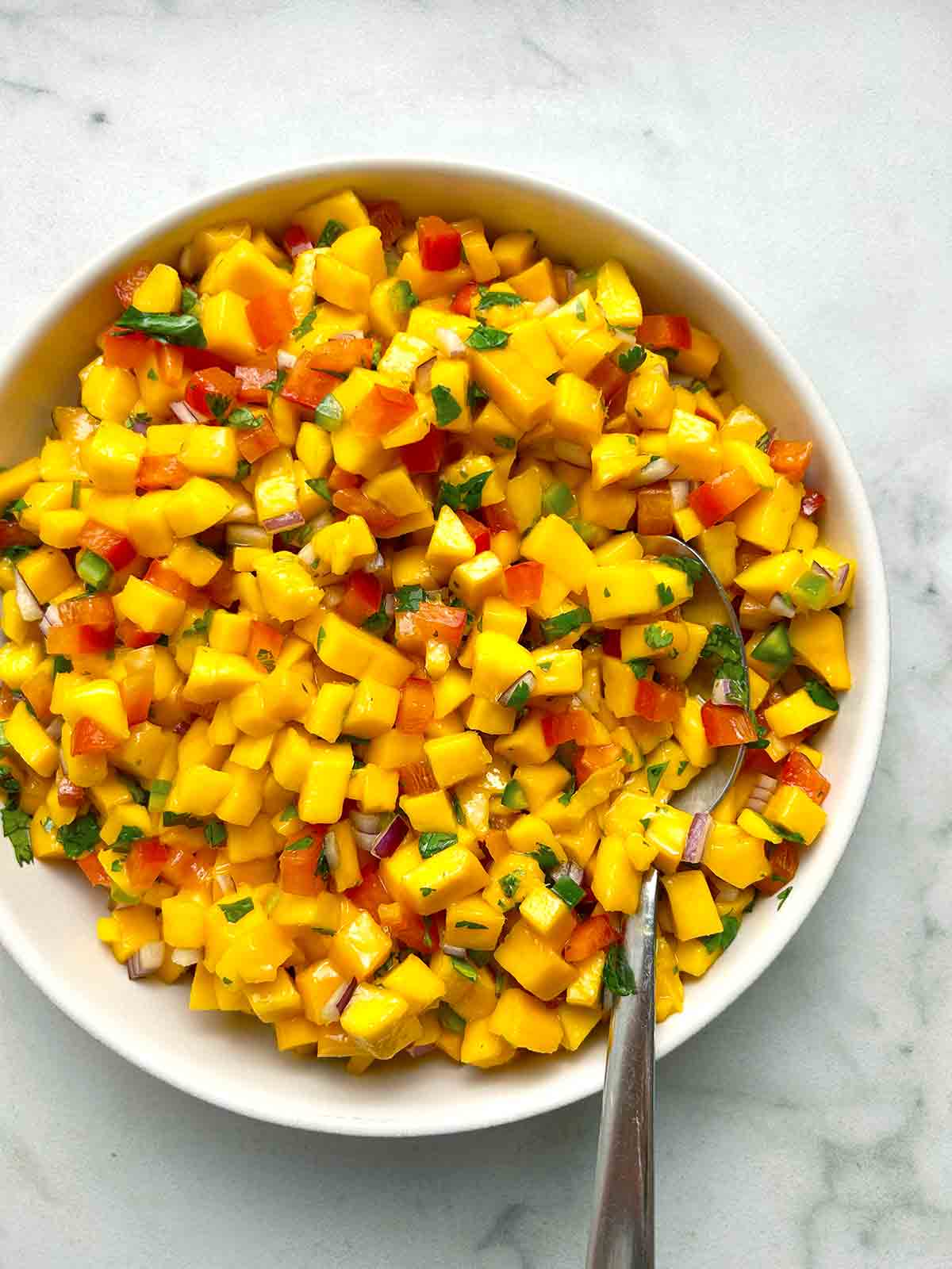 A bowl of mango salsa with diced mango, red onion, and cilantro, mixed with red bell pepper. A spoon rests in the bowl.