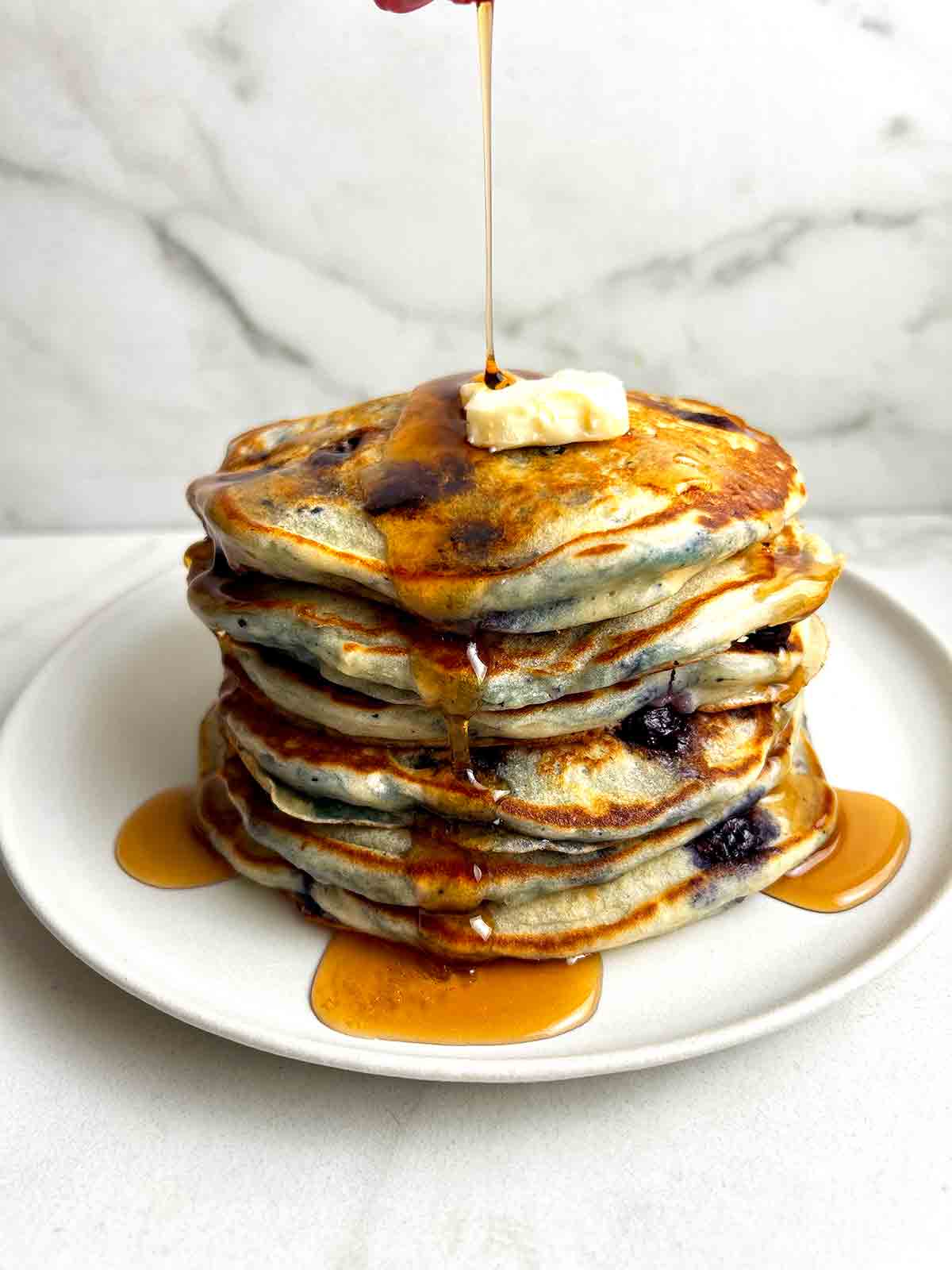 syrup being poured over stack of blueberry pancakes.